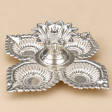 Front side angle view of a German Silver Pooja Diya showing the raised central floral holder, four surrounding decorative leaf shaped diya cups, sculpted silver borders and compact elevated construction, photographed on a warm beige background to clearly display the product depth, height and traditional design for pooja ceremonies, Diwali decoration, festivals and gifting.