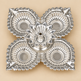 Top view of a German Silver Pooja Diya showing the symmetrical four cup arrangement around the central flower shaped holder, with each leaf shaped diya cup featuring detailed ridged interiors, ornamental scalloped borders and a bright polished silver finish, photographed on a clean beige studio background for a clear view of the complete traditional design and layout.