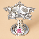 Slightly top-angled product image of a German Silver Diya placed on a premium beige studio background, clearly showing the interior of the square floral bowl, textured reflective finish inside the diya, pointed corner extensions, decorative beadwork along the rim, ringed pedestal stem and broad round base, allowing customers to view the diya from another angle and better understand its bowl shape, decorative styling and traditional devotional use.