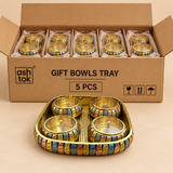 Five gift bowls tray sets shown as a bulk quantity variant with simple brown corrugated cardboard packaging on a warm beige studio background, featuring coordinated rectangular golden trays with rounded corners and matching sets of four round lidded bowls with multicolor patterned outer bands, polished golden lids and small top knobs. The packaging is practical and non-premium, with multiple sets arranged safely inside the cardboard box and one complete tray set displayed clearly in front, making the image 