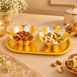 Gold and Silver Plated Dry Fruit Serving Set beautifully arranged on an elegant beige dining table, featuring two decorative floral bowls filled with almonds, cashews, and pistachios. Each bowl features alternating gold and silver engraved flower petals and a matching ornamental gold serving spoon, while the polished oval tray displays a raised beaded border and delicate floral detailing. Surrounded by tasteful table decorations and warm ambient lighting, the serving set creates an attractive presentation f