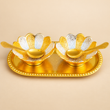 Angled product view of an elegant Gold and Silver Plated Dry Fruit Bowl Set displaying two decorative flower-petal bowls arranged on a polished oval golden tray. Each bowl features alternating silver and gold engraved petals with intricate floral and leafy patterns, raised bases, and matching gold-toned serving spoons. The tray features delicate surface engraving and a rounded beaded rim, creating an attractive serving arrangement for almonds, cashews, pistachios, sweets, and festive snacks.