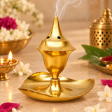 Golden brass agarbati flower incense holder shown in use in a warm devotional setting with soft incense smoke rising from the pointed top, surrounded by tasteful flowers, brass puja accessories and a gently lit diya on a beige-toned surface, demonstrating practical use during daily worship, meditation, festive rituals and home mandir ceremonies without any deity idol included in the scene.
