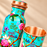 Detailed close-up of a green enamel printed pure copper water bottle and matching drinking glass highlighting the turquoise-green enamel surface, pink and white flowers, decorative brown branches, green leaves, copper-coloured screw cap and polished copper glass interior. The image showcases the colourful botanical print, smooth metallic rim and intricate cap detailing against a beige studio background.