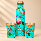 Angled product view of a green enamel printed pure copper water bottle with two matching glasses, one standing upright and the other placed sideways to reveal its polished copper interior. The turquoise floral exterior features pink flowers, green foliage and brown branches, complemented by a copper-coloured bottle cap. Displayed on a beige background to highlight the matching drinkware design.