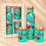 Green enamel printed pure copper water bottle with two matching floral drinking glasses displayed alongside a simple cream-coloured cardboard gift box with gold floral patterns and a transparent front window. The turquoise-green drinkware features pink flowers, decorative branches, green leaves and polished copper interiors. Suitable for housewarming presents, wedding gifts, festive gifting and traditional return gifts.