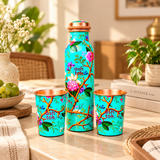 Green enamel printed pure copper water bottle with two matching drinking glasses arranged on a beige dining table in a bright home interior. The turquoise-green bottle features delicate pink and white flowers, green leaves, brown branches and a copper-coloured cap. Both glasses have matching botanical prints and polished copper interiors, suitable for everyday drinking water and tableware decoration.