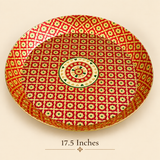 Studio photograph of a circular 17.5-inch brass decorative tray with a vibrant red and gold patterned surface and raised edges. Set on a warm beige background with '17.5 Inches' text and a small floral motif displayed underneath.
