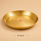 Studio product shot of a 16-inch round brass puja plate featuring a concentric hammered dimple pattern across its interior base and raised rim. Set against a warm beige backdrop with '16 Inch' text displayed beneath and an Ashtok logo watermark at the center of the plate.