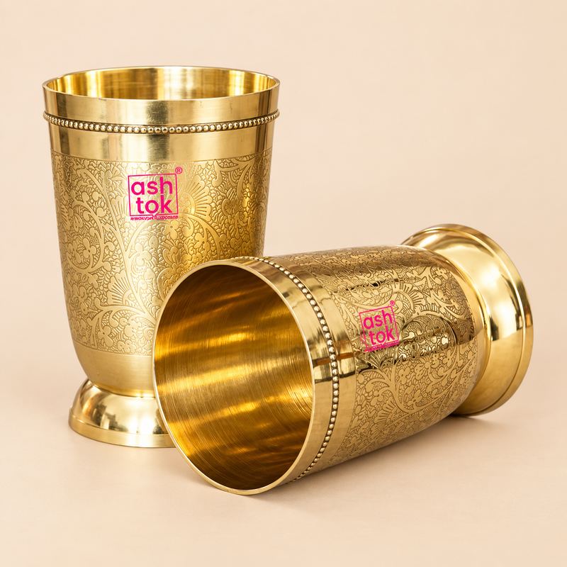 Handcrafted brass water glass set featuring two engraved brass glasses with a polished golden finish, floral etched design, smooth brass interior and sturdy flared base, displayed on a premium beige studio background for elegant product presentation.