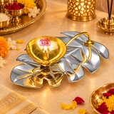 Beautifully illuminated Leaf Design Brass Puja Diya displayed in an elegant traditional pooja setting, featuring a polished golden brass oil lamp bowl with a glowing cotton wick flame mounted on a decorative silver-finish metallic leaf-shaped tray. The circular brass bowl has a reflective golden interior with concentric detailing and a central cylindrical wick holder supporting the burning flame. A gracefully curved golden metallic stem extends over the leaf arrangement, connecting the golden bowl.