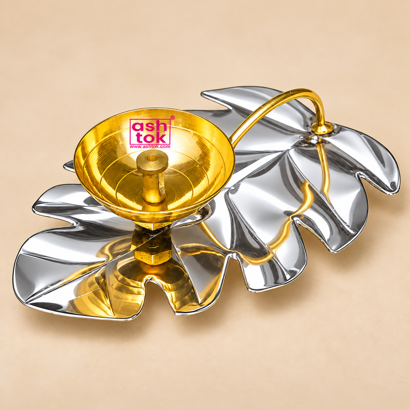 Elegant Leaf Design Brass Puja Diya displayed on a premium beige studio background, featuring a beautifully polished golden brass oil lamp bowl mounted on a decorative silver-coloured metallic leaf-shaped tray. The diya features a wide circular brass bowl with smooth rounded edges, a reflective golden interior, fine concentric circular detailing, and a centrally positioned cylindrical wick holder with a small opening for a cotton wick. A graceful curved golden brass stem extends from the bowl towards the op