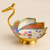 Meenakari duck design fruit bowl shown in an elegant festive lifestyle setting filled with assorted dry fruits and nuts including almonds, cashews, pistachios, raisins and dates. The decorative bowl features a polished golden duck-shaped neck and head, colorful feather-inspired panels with intricate traditional Meenakari floral artwork and a stable raised pedestal base.