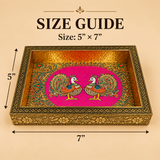 Comprehensive size guide diagram for a rectangular Meenakari peacock decorative wooden serving tray showing exact dimensions of 5 inches in width and 7 inches in length with measurement arrows on a warm beige background. The tray features a vibrant pink base with colorful dual peacock artwork, a hammered gold interior rim, and an engraved floral pattern along the outer brass frame.