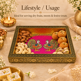 A high-angle festive lifestyle setup of the Meenakari peacock serving tray filled with Indian sweets like motichoor laddu and dry fruits including almonds, cashews, and pistachios. Set on a marble surface decorated with a glowing brass diya, fresh white jasmine flowers, and Kaju Katli sweets in a golden bowl, perfect for festive gifting and entertaining.