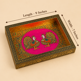 Dimension diagram of a rectangular handcrafted wooden serving tray featuring a pair of royal Meenakari peacocks on a bright magenta-pink circular background surrounded by colorful floral motifs. Black measurement arrows indicate dimensions of 9 inches in length along the top and 7 inches in width along the side. The tray features a hammered gold inner wall finish and an engraved gold floral outer border.