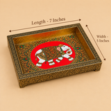 Dimension diagram of a rectangular handcrafted wooden tray featuring a central royal elephant Meenakari artwork against a bright red circular background surrounded by ornate yellow and green floral patterns. Black measurement arrows indicate dimensions of 7 inches in length and 5 inches in width across the top rim. The tray is bordered with an intricate, dark gold and black floral engraved frame resting on a soft beige background.
