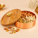 Traditional copper-coloured metal dry fruit bowl displayed open in an elegant festive setting, filled with assorted almonds, cashews, pistachios and golden raisins. Its natural wooden lid with a decorative golden leaf-shaped handle rests partially beside the container, revealing the spacious interior and smooth copper-toned base. The hammered exterior, natural wood grain and warm beige surroundings create an attractive presentation suitable for Diwali celebrations, wedding gifting, housewarming occasions.