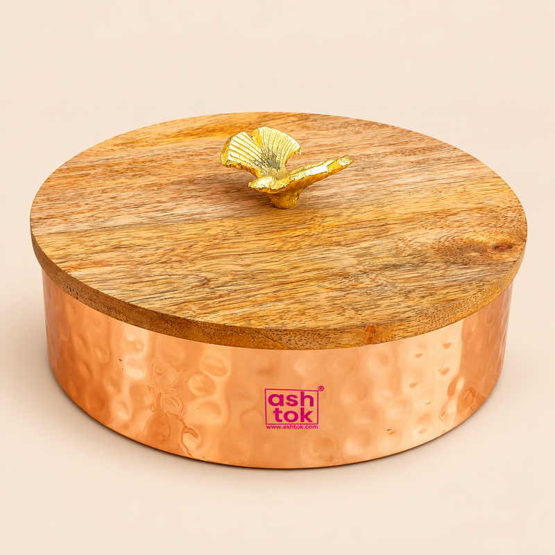 Traditional metal dry fruit bowl featuring a warm copper-coloured hammered finish, round cylindrical body with decorative circular impressions, and a natural wooden lid topped with an elegant golden leaf-shaped handle. The compact storage container measures approximately 4 inches in diameter and 3 inches in height, making it suitable for storing assorted dry fruits, sweets, chocolates and small household essentials. Displayed on a beige studio background, this decorative lidded bowl is ideal for festive.