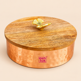 Traditional metal dry fruit bowl featuring a warm copper-coloured hammered finish, round cylindrical body with decorative circular impressions, and a natural wooden lid topped with an elegant golden leaf-shaped handle. The compact storage container measures approximately 4 inches in diameter and 3 inches in height, making it suitable for storing assorted dry fruits, sweets, chocolates and small household essentials. Displayed on a beige studio background, this decorative lidded bowl is ideal for festive.