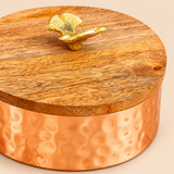 Detailed close-up photograph of a decorative copper-coloured metal dry fruit box highlighting the natural wood grain texture of its circular wooden lid, beautifully crafted golden leaf-shaped handle and hammered circular impressions on the copper-toned container body. The image showcases the contrast between the warm wooden surface, reflective metallic finish and ornamental lid handle, creating a traditional yet elegant storage container suitable for festive gifts, decorative displays and dry fruit.