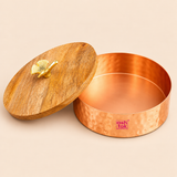 Open view of a decorative copper-coloured metal dry fruit storage bowl showcasing its spacious circular interior, smooth copper-toned base, straight cylindrical walls and hammered exterior texture. The removable natural wooden lid is positioned beside the container, displaying its distinctive wood grain pattern and golden decorative leaf-shaped handle. The photograph highlights the practical storage space and contrasting wooden and metallic finishes, suitable for keeping almonds, cashews, pistachios, raisin