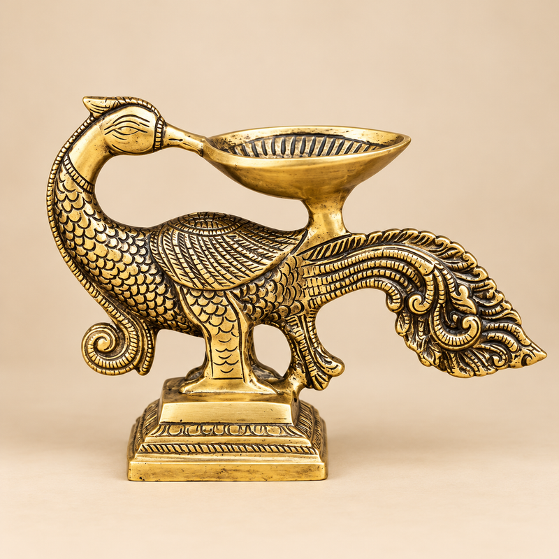 Traditional antique brass peacock diya featuring an intricately sculpted peacock with an elegantly curved neck, detailed feather engravings, decorative wings and an elaborate flowing tail. A shallow brass oil lamp bowl is positioned above the peacock's back and extends towards its beak, creating a distinctive traditional deepam design. The entire structure rests on an ornamental rectangular pedestal with engraved borders