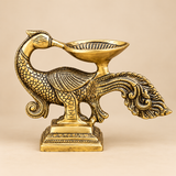 Traditional antique brass peacock diya featuring an intricately sculpted peacock with an elegantly curved neck, detailed feather engravings, decorative wings and an elaborate flowing tail. A shallow brass oil lamp bowl is positioned above the peacock's back and extends towards its beak, creating a distinctive traditional deepam design. The entire structure rests on an ornamental rectangular pedestal with engraved borders