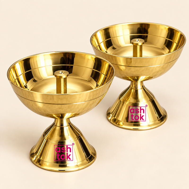 Studio product image showcasing a pair of shiny polished brass pedestal oil lamps (diyas) with central wick holders and flared footed bases, set against a plain beige backdrop with Ashtok logo watermarks.