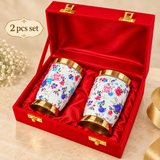 Set of 2 floral printed brass water glasses presented in a red gift box, featuring polished golden interiors, decorative floral exteriors and gold finished rims and bases, suitable for weddings, festivals and special occasion gifting.