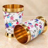 Printed brass water glasses with colorful floral design, polished golden interior, decorative gold rim and sturdy base, presented as a matching pair on a premium beige studio background for elegant drink serving and special occasions.
