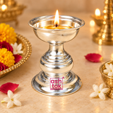 Traditional silver finish Puja Deepam shown in a warm devotional lifestyle setting with a lit wick and gentle flame in the upper bowl, surrounded by tasteful flowers and festive decorative elements on a beige-toned surface, demonstrating its use for daily puja, diya lighting, prayer rituals, Diwali celebrations, home temple ceremonies, festive worship and traditional Indian religious occasions without any deity idol in the scene.
