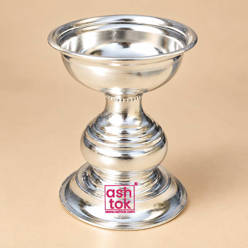 Polished silver finish Puja Deepam with a traditional pedestal design, wide shallow top bowl, rounded rolled rim, decorative narrow neck, bulbous central body with layered ring detailing and a broad stable circular base, photographed as a clean front-facing product image on a premium beige studio background for daily puja, festive worship, home temple use, pooja room setup, religious ceremonies, return gifting and traditional Indian devotional occasions.