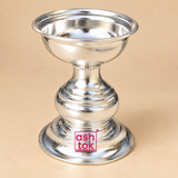 Traditional silver finish Puja Deepam shown from a right three-quarter angle with a reflective shallow oil bowl, smooth rolled rim, decorative neck detailing, rounded stepped central body and wide flared base, photographed on a clean beige studio background to clearly display the shape, depth, polished metal surface and traditional craftsmanship for home puja, pooja rituals, festive lighting and religious use.