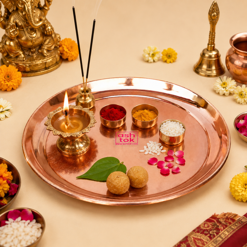 Copper puja thali arranged with a lit brass diya, kumkum, turmeric, rice, sweets, incense, and decorative flowers next to a Lord Ganesha idol.