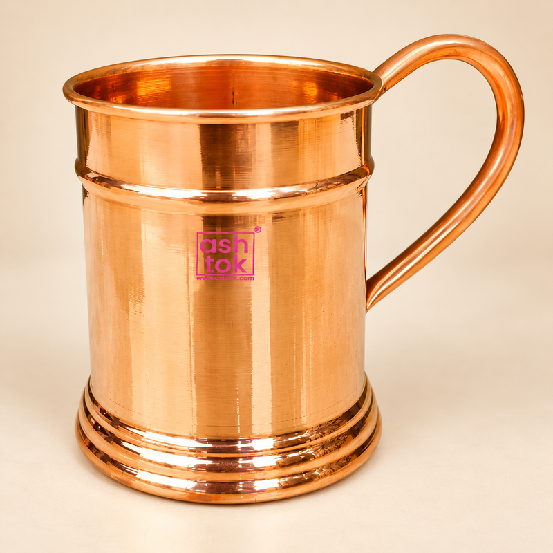 Pure copper beer mug with a polished copper finish, cylindrical body, rounded handle, rolled top rim and decorative stepped base, professionally presented on a premium beige studio background for cocktails, Moscow Mule drinks and beverage serving.