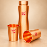 Angled view of a pure copper water bottle with two matching copper glasses, one standing upright and the other positioned sideways to reveal its polished copper interior. The bottle features a tall curved body, smooth brushed copper finish and screw cap. Displayed against a clean beige background to highlight the traditional copper drinkware set.
