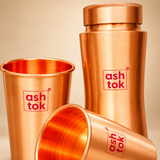 Detailed close-up of a pure copper water bottle and matching drinking glasses highlighting the brushed satin copper finish, curved bottle shape, textured screw cap, smooth glass rims and polished copper interiors. The close-up shows the natural copper colour and metallic surface detailing against a warm beige background.