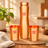 Pure copper water bottle with two matching copper drinking glasses arranged on a wooden dining table in a warm home interior. The drinkware features a satin-brushed copper finish, gently curved bottle shape and polished copper glasses, surrounded by subtle home accessories and natural lighting. Suitable for serving water at home, family dining and everyday hydration.