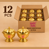 welve polished brass sindoor boxes presented in a simple brown corrugated cardboard package with internal compartments, with two full brass sindoor boxes placed in front for a clear view of the decorative petal-shaped lids, rounded bowls, small top knobs, polished golden surface and stable pedestal bases, photographed on a warm beige background to demonstrate a practical 12 piece pack for bulk orders, festive gifting, pooja accessories, wedding return gifts, religious events and wholesale supply without lux
