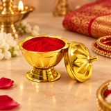 Traditional golden brass sindoor box displayed in an elegant Indian wedding setting with its decorative floral lid removed and placed beside the container. The bowl is filled with bright red sindoor powder and positioned on a beige marble surface, surrounded by jasmine flowers, red rose petals, bridal bangles, traditional brass lamps and festive fabric. The image illustrates its use during Hindu marriage ceremonies, sindoor rituals, daily puja and traditional wedding celebrations.