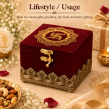 High-angle lifestyle product presentation of a closed royal maroon velvet jewellery box placed on a festive table layout surrounded by delicate white baby's breath flowers, fresh red rose petals, a small golden bowl filled with mixed dry fruits like almonds, cashews, and pistachios, a glowing tea light candle, and wrapped gift boxes, demonstrating its use for Indian wedding return gifts, jewellery storage, dry fruit packaging, and festive celebrations.