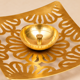 Detailed close-up image of a Square Shape Brass Jali Patta Diya focusing on the polished brass center oil bowl, sturdy upright wick holder and surrounding floral and leaf-inspired jali cutwork, with the curved brass plate edges and fine metal surface texture clearly visible against a premium beige studio background, designed to showcase the craftsmanship, cutout detailing, brass finish and traditional decorative styling of the diya.