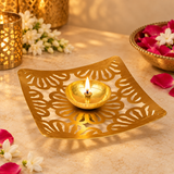 Square Shape Brass Jali Patta Diya shown in a warm festive setting with a small lit flame in the central brass oil cup, surrounded by tasteful white flowers, pink rose petals and subtle brass decorative accessories on a beige surface, demonstrating practical use for daily worship, Diwali lighting, festive puja, evening aarti, home mandir rituals and traditional decorative settings without any idol or deity figure included.