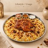 A top-angle lifestyle shot featuring a large, round stainless steel serving platter (mandi platter) filled with flavorful saffron basmati rice topped with a roasted whole chicken, fried cashews, raisins, fried onions, and star anise. Three small stainless steel bowls containing side dishes—fresh cucumber-tomato salad, gravy, and raita yogurt dip—are set on the upper rim inside the platter. The background is elegantly styled in a warm, rustic aesthetic with a brass lantern on the left, a decorative ceramic v
