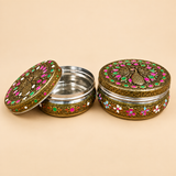 Studio display photograph of two Stainless Steel Meenakari Puri Boxes on a neutral tan background. The container on the left sits open with its decorated lid tilted beside the food-grade stainless steel interior, while the container on the right is closed to show off the intricate embossed gold peacock lid with pink and green Meenakari enamel and floral side border detailing.
